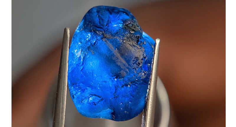 Close-up of a raw deep blue sapphire gemstone held by metal tweezers, representing the September birthstones sapphire.
