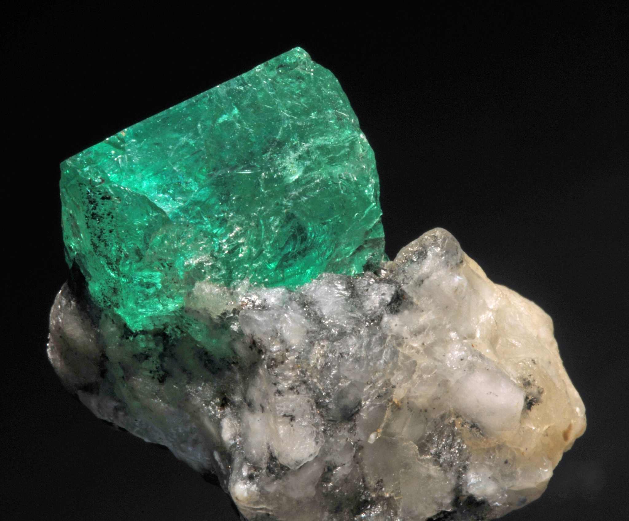 Raw bright green emerald birthstone crystal on rough white and gray mineral matrix against black background