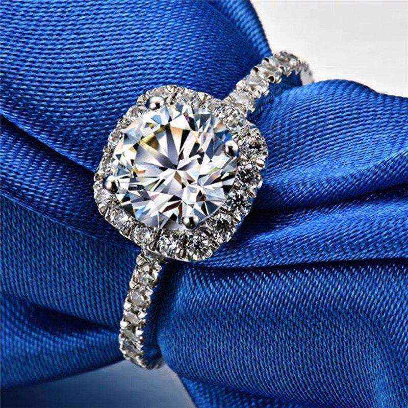 Close-up of a 1.5 CTW colorless round brilliant cut moissanite halo engagement ring in sterling silver with pavé accent stones on a blue fabric background