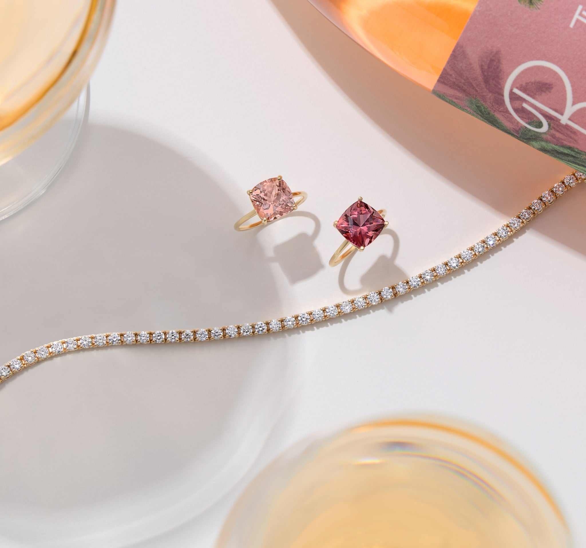 Two cushion-cut pink tourmaline rings set in 14k gold displayed with a diamond tennis bracelet on a white surface.