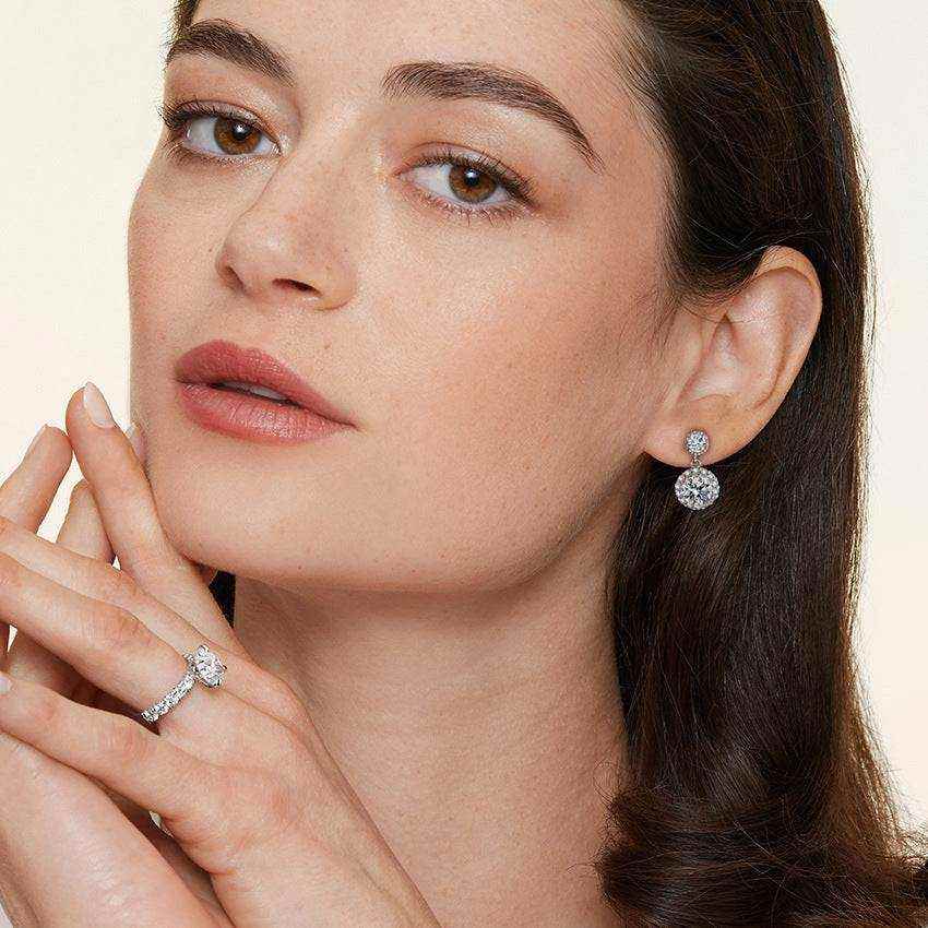 Woman wearing 1 CTW DEW colorless round cut moissanite halo drop earrings in 925 sterling silver with push back studs.