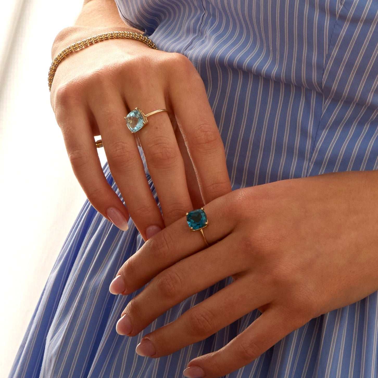 Hands wearing 14k gold rings featuring cushion-cut sky light blue topaz gemstones on a person in a blue striped dress