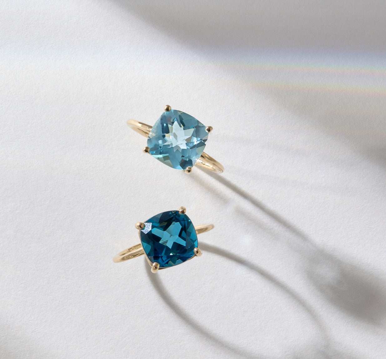 Two gold rings featuring cushion cut sky light blue topaz gemstones on a white surface with soft shadows
