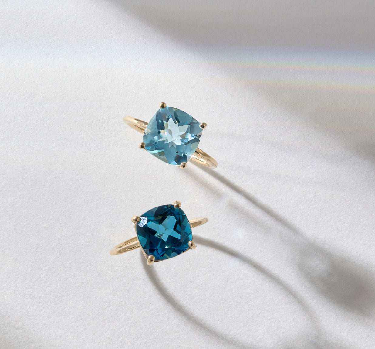 Two 14k gold rings featuring cushion cut blue topaz gemstones in different shades, displayed on a light surface with soft shadows.
