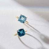 Two 14k gold rings featuring cushion cut blue topaz gemstones in different shades, displayed on a light surface with soft shadows.