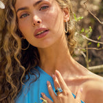 Woman wearing a sky light blue topaz ring with cushion cut gemstone and gold band, styled with hoop earrings and blue top.