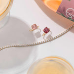 Two cushion-cut pink tourmaline rings set in 14k gold displayed with a diamond tennis bracelet on a white surface.