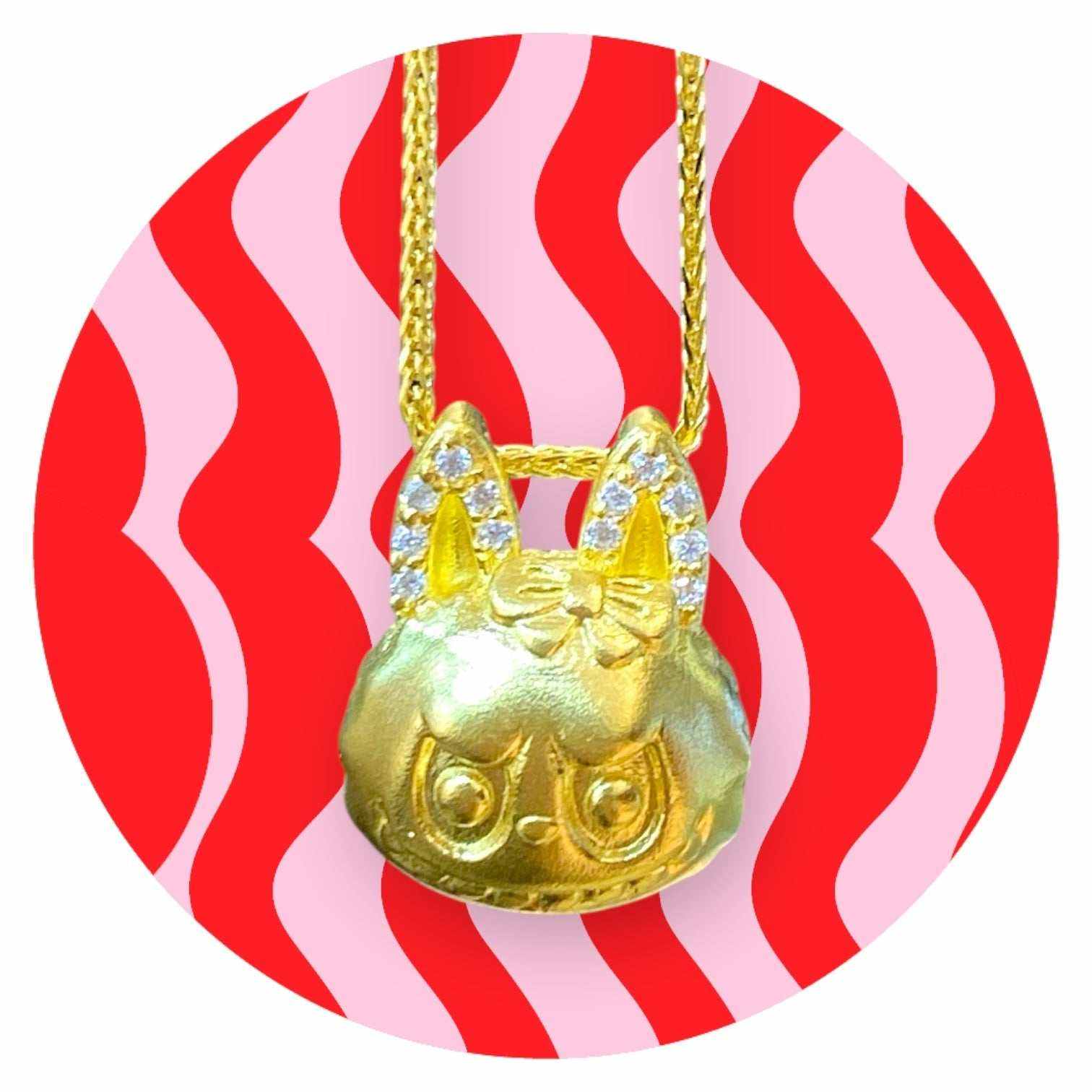 24K gold Labubu-inspired doll-head pendant with sparkling moissanite stones on a gold chain against red and pink wavy background
